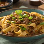 A bowl of Mongolian ground beef noodles topped with green onions and sesame seeds, served on a wooden table with chopsticks and side vegetables.